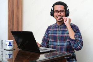 Adult Asian man sitting in front of laptop smiling while touching his headset mic